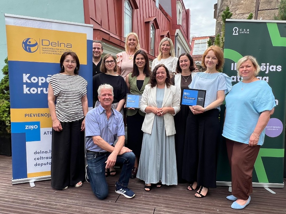 Pasākuma dalībnieki kopīgā grupas foto āra terasē starp “Delna” un “CSR Latvia” stendiem. Daļa dalībnieku tur rokās atzinības plāksnes, kas saistītas ar pasākumā pasniegtu novērtējumu vai apbalvojumu.