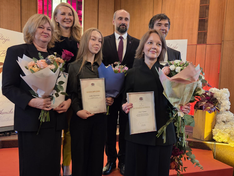 Grupas fotogrāfija apbalvošanas pasākumā. Priekšplānā trīs sievietes ar ziediem un atzinības rakstiem; otrajā rindā redzami trīs cilvēki, tostarp ministre, valsts sekretārs un vēl viens kolēģis.