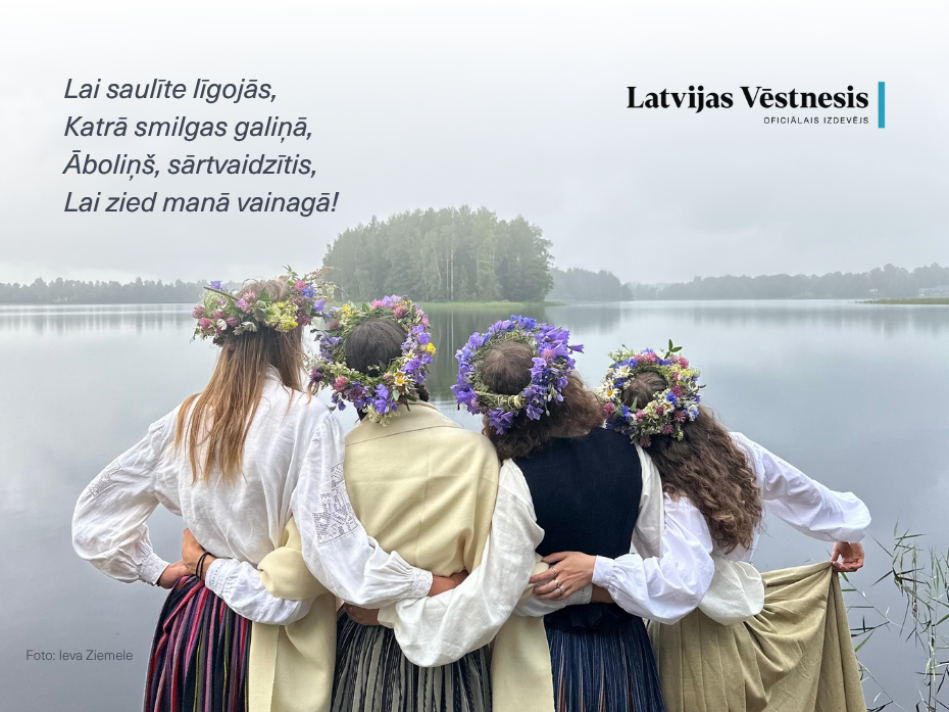 Četras sievietes tautastērpos ar Jāņu vainagiem stāv kopā, apskāvušās, pie ūdenstilpes, svinot vasaras saulgriežus. Uz attēla "Latvijas Vēstneša" apsveikums.